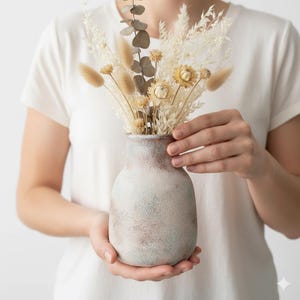 May include: A ceramic vase with a textured, mottled finish in shades of white, brown, and blue holds a bouquet of dried flowers. The arrangement includes fluffy beige bunny tails, yellow daisies, and brown eucalyptus leaves. The vase is held by a person.