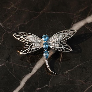 May include: A silver dragonfly brooch with intricate wing detailing. The wings are adorned with small, clear crystals and the body features three blue gemstones. The brooch is set against a dark, marbled surface.