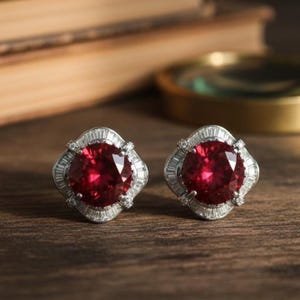 May include: A pair of stud earrings featuring large, round, deep red gemstones surrounded by a halo of smaller, clear stones. The earrings are set in a silver-colored metal and rest on a wooden surface.