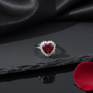 Może przedstawiać: Srebrny pierścionek z dużym, czerwonym kamieniem szlachetnym w kształcie serca. Kamień otoczony jest aureolą mniejszych, przezroczystych kamieni. Pierścionek prezentowany jest na ciemnej, teksturowanej powierzchni, z czerwonym płatkiem w rogu.