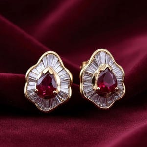 May include: A pair of gold-toned stud earrings. Each earring features a teardrop-shaped, deep red gemstone at the center, surrounded by a halo of baguette-cut, clear gemstones. The earrings are set against a deep red velvet background.