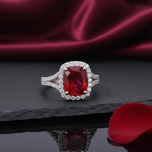 Może przedstawiać: Srebrny pierścionek z dużym, czerwonym kamieniem szlachetnym o szlifie poduszkowym, otoczonym aureolą mniejszych, przezroczystych kamieni. Pierścionek ma rozszczepioną szynę i jest prezentowany na ciemnej powierzchni.