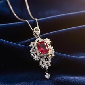 May include: A silver necklace with a pendant featuring a large, rectangular, red gemstone. The gemstone is surrounded by a halo of smaller, clear gemstones and decorative filigree. A small, round gemstone dangles from the bottom of the pendant.
