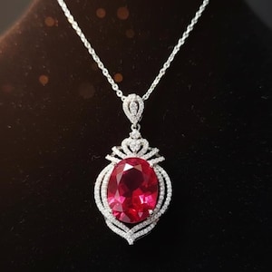 May include: A silver necklace with a large, oval, ruby-red gemstone pendant. The pendant is surrounded by a halo of small, clear gemstones. The necklace hangs on a delicate silver chain, creating a luxurious and elegant look.