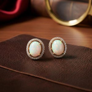 Könnte beinhalten: Ovale Opal-Ohrstecker mit einem Kranz aus kleinen, klaren Edelsteinen in Gold gefasst. Die Opale haben eine milchig-weiße Farbe mit subtilem Farbspiel. Die Ohrringe werden auf einer braunen Lederoberfläche präsentiert.