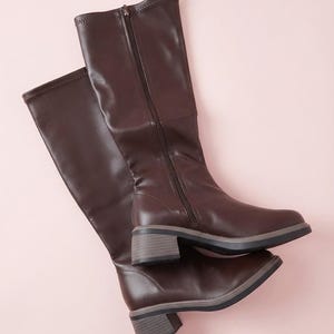 Peut inclure: Une paire de bottes marron foncé arrivant au genou avec une finition lisse, une fermeture éclair latérale et un talon bloc à base grise. Les bottes présentent un bout arrondi et une tige structurée arrivant sous le genou, offrant une silhouette épurée.