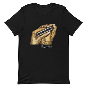 May include: Black t-shirt featuring a hand-drawn illustration of a harmonica held in two hands. The text "Truancy Point" is written below the hands in a handwritten font. The design is in shades of brown, white, and black.