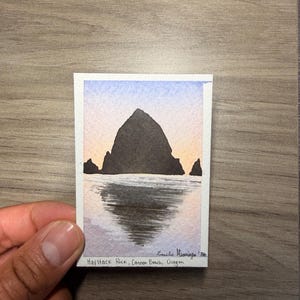 Peut inclure: Une petite peinture rectangulaire à l'aquarelle représentant Haystack Rock à Cannon Beach, dans l'Oregon. L'œuvre présente une silhouette sombre de la formation rocheuse sur un ciel pastel doux, avec un reflet sur l'eau. Le texte indique Haystack Rock, Cannon Beach, Oregon.
