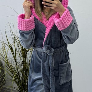 May include: A gray hooded robe with pink trim on the hood and cuffs. The robe has a matching belt and pockets. The soft, plush material is visible. The robe is designed for comfort and relaxation.