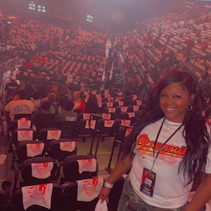 May include: A white t-shirt with red and orange text reading Dreamville, worn with camouflage cargo trousers and a black lanyard featuring a rectangular pass that reads The Fall Off, standing in a large indoor arena with rows of black seats.