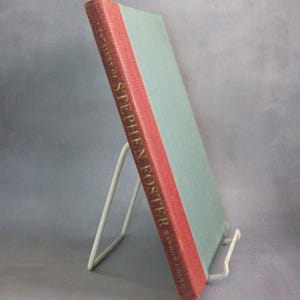 May include: A book titled "A Treasury of Stephen Foster" with a light blue cover and a red spine. The book is propped up on a white metal stand against a gray background.