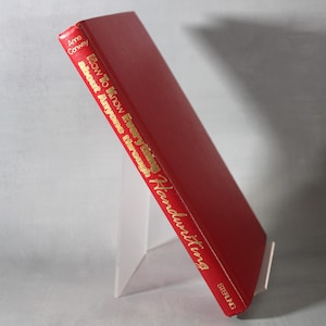 Op de afbeelding: Een rood hardcover boek getiteld "How to Know Everything about Anyone through Handwriting" door Anne Conway. Het boek wordt getoond op een witte standaard, met gouden letters op de rug. De kaft van het boek is effen dieprood.