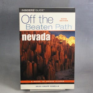 May include: A travel guide titled "Off the Beaten Path Nevada" with a Fifth Edition label. The cover features a photograph of a desert landscape with tall, reddish-brown rock formations. The guide is by Heidi Knapp Rinella.