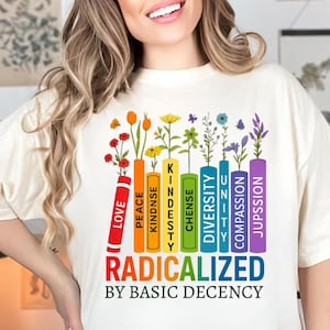 May include: Cream-colored t-shirt with a colorful graphic. The graphic features a row of books with words like "Love," "Peace," and "Diversity," each with flowers sprouting from the top. Below, the text reads "Radicalized by Basic Decency."