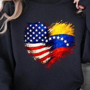 May include: A navy blue sweatshirt featuring a heart-shaped design. The left side of the heart displays the American flag, while the right side showcases the Venezuelan flag. The flags are rendered with a brushstroke effect.