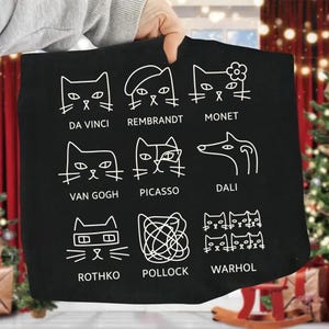 May include: Black tote bag featuring white line drawings of cats in the style of famous artists, including Da Vinci, Rembrandt, Monet, Van Gogh, Picasso, Dali, Rothko, Pollock, and Warhol. The artist's name is under each cat.