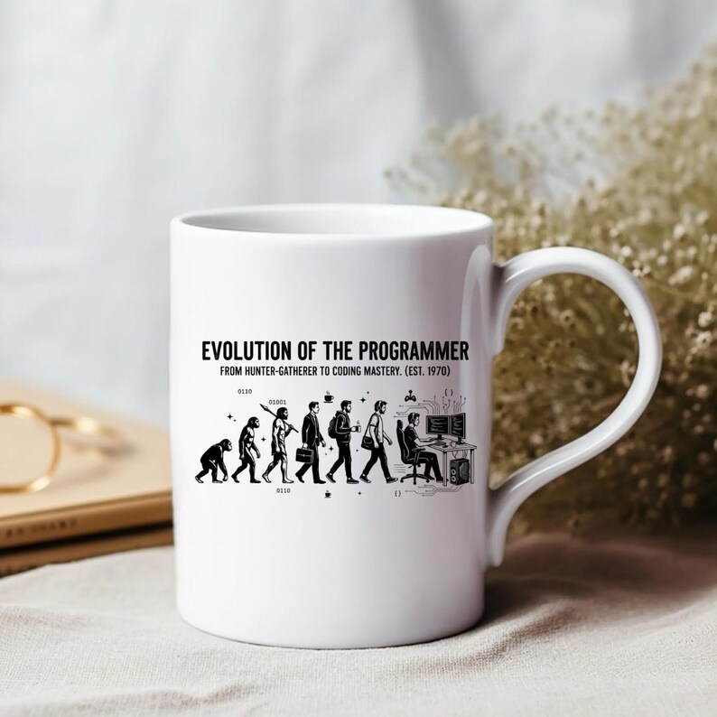 Op de afbeelding: Een witte keramische mok met een zwarte afbeelding die de evolutie van mens naar programmeur weergeeft. De tekst luidt: EVOLUTION OF THE PROGRAMMER FROM HUNTER-GATHERER TO CODING MASTERY. (EST. 1970). Het ontwerp toont figuren in een proces van ontwikkeling.