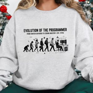 Op de afbeelding: Een lichtgrijs sweatshirt met ronde hals en een zwarte grafische print met de titel EVOLUTION OF THE PROGRAMMER FROM HUNTER-GATHERER TO CODING MASTERY. (EST. 1970). Het ontwerp toont de menselijke evolutie die eindigt bij een persoon achter een computer.