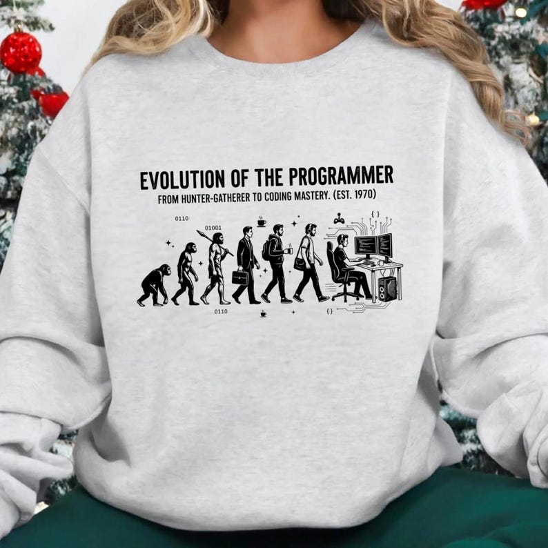 Op de afbeelding: Een lichtgrijs sweatshirt met ronde hals en een zwarte grafische print met de titel EVOLUTION OF THE PROGRAMMER FROM HUNTER-GATHERER TO CODING MASTERY. (EST. 1970). Het ontwerp toont de menselijke evolutie die eindigt bij een persoon achter een computer.