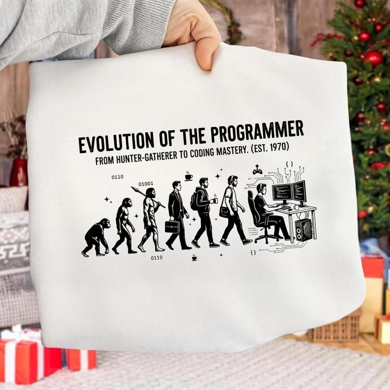 Op de afbeelding: Een wit sweatshirt met een zwarte afbeelding met de titel EVOLUTION OF THE PROGRAMMER FROM HUNTER-GATHERER TO CODING MASTERY. EST. 1970. Het ontwerp toont de evolutie van een primaat naar een persoon achter een computerbureau met binaire code.
