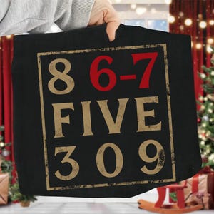 May include: Black tote bag with a rectangular design featuring the numbers 8, 6-7, FIVE, 309 in a distressed, vintage style. The numbers are in a beige color, with the 6-7 in red. The design is framed by a thin, beige border.
