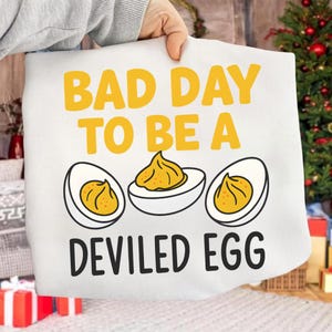 Puede incluir: Bolso de mano blanco con el texto "BAD DAY TO BE A DEVILED EGG" en amarillo y negro. El diseño incluye tres huevos rellenos con relleno amarillo.
