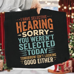 Puede incluir: Un artículo de tela negro con el texto I HAVE SELECTIVE HEARING SORRY YOU WEREN'T SELECTED TODAY TOMORROW ISN'T LOOKING GOOD EITHER en letras naranjas, azul claro y amarillas con líneas horizontales y pequeñas estrellas.