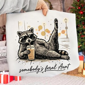 Puede incluir: Una sudadera blanca con el gráfico de un mapache con gafas de sol, tumbado mientras sostiene un vaso de café con una pata levantada. El diseño incluye farolas y el texto "somebody's feral Aunt" en una fuente cursiva de color negro.