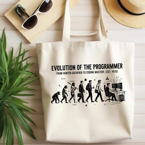 Op de afbeelding: Een cr&egrave;mekleurige canvas tas met een zwarte grafische print met de titel EVOLUTION OF THE PROGRAMMER FROM HUNTER-GATHERER TO CODING MASTERY. (EST. 1970). De illustratie toont de evolutie van een primaat naar een computerprogrammeur.