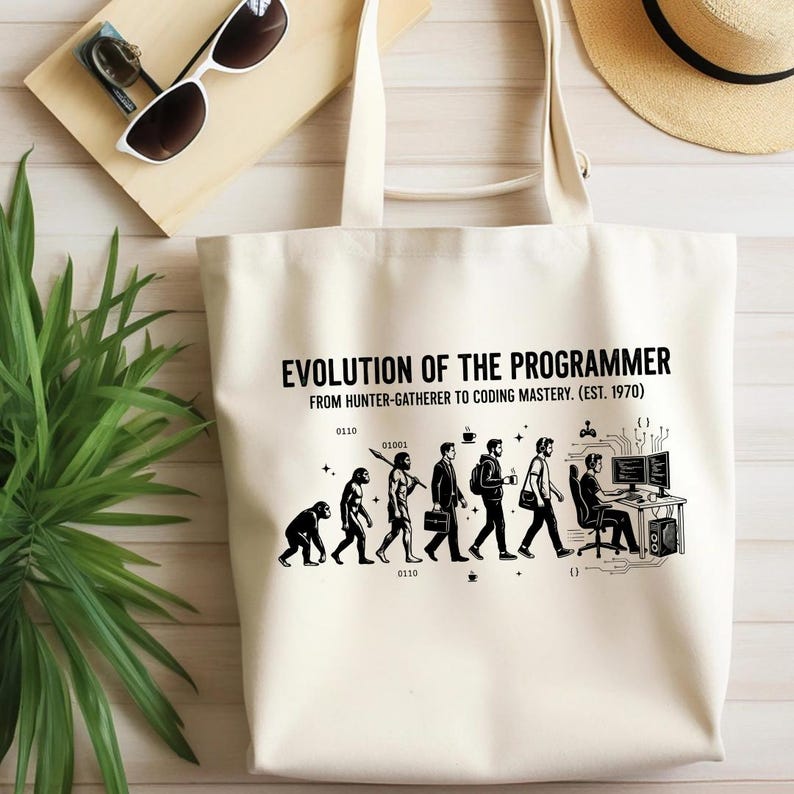 Op de afbeelding: Een cr&egrave;mekleurige canvas tas met een zwarte grafische print met de titel EVOLUTION OF THE PROGRAMMER FROM HUNTER-GATHERER TO CODING MASTERY. (EST. 1970). De illustratie toont de evolutie van een primaat naar een computerprogrammeur.