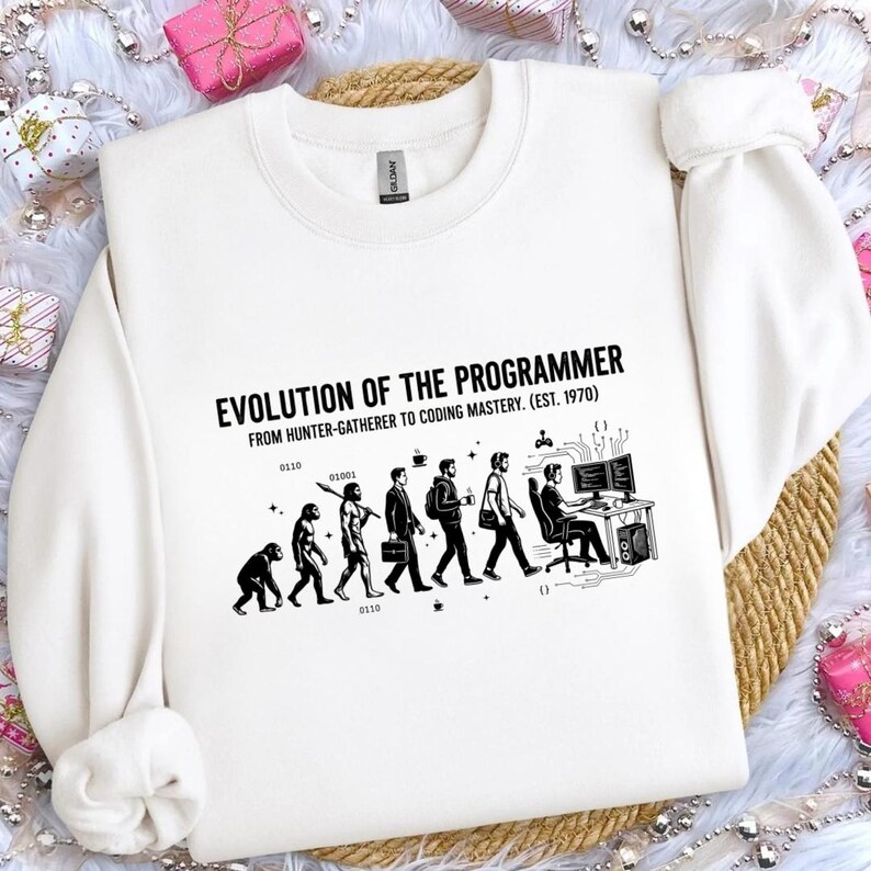 Op de afbeelding: Een cr&egrave;mekleurige sweater met ronde hals en een zwarte grafische print met de tekst EVOLUTION OF THE PROGRAMMER FROM HUNTER-GATHERER TO CODING MASTERY. EST. 1970. Het ontwerp toont de evolutie van primaat naar een persoon achter een computer.