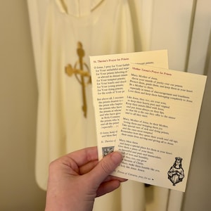 Könnte beinhalten: Eine Hand hält zwei Gebetskarten mit religiösem Text und einer kleinen Illustration. Die Karten sind cremefarben mit schwarzem Text. Im Hintergrund ist ein weißes religiöses Gewand mit einem goldenen Kreuz zu sehen.