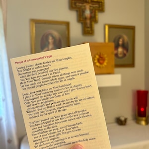 Könnte beinhalten: Religiöse Szene mit einer Gebetskarte mit Text, die vor einem Regal mit gerahmter religiöser Kunst und einem goldenen Kruzifix gehalten wird. Der Text der Karte ist sichtbar, und der Hintergrund umfasst eine rote Kerze.