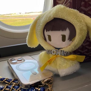 May include: A plush doll with a yellow bunny-eared hood and a rhinestone collar that says "WACHINE". A clear phone case with a gold chain strap and a blue patterned scarf is also visible.