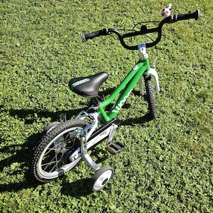 Op de afbeelding: Een groene en witte kinderfiets met zijwieltjes, een zwart zadel en zwart stuur. De fiets staat op een groene grasachtige ondergrond. De fiets heeft een bel op het stuur.