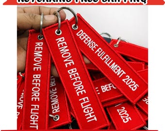 May include: A collection of rectangular red fabric keychains with white embroidered text. Some keychains feature the text REMOVE BEFORE FLIGHT, while others display DEFENSE FULFILLMENT 2025. Each keychain includes a silver metal keyring.