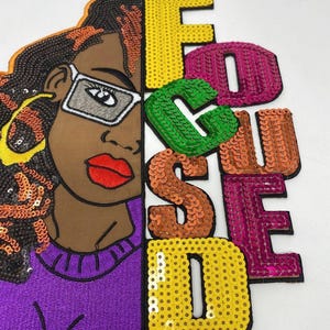 Könnte beinhalten: Gestickter Aufnäher mit dem Gesicht einer Frau mit Sonnenbrille und dem Wort "FOCUSED" in Paillettenbuchstaben. Die Frau hat braune Haut, rote Lippen und einen lila Pullover. Die Buchstaben sind gelb, grün, orange und pink.