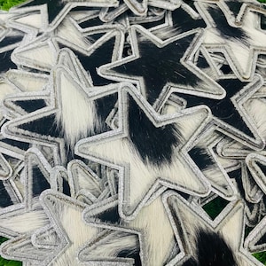 Könnte beinhalten: Ein Haufen sternförmiger Aufnäher mit silberner Glitzerborte. Die Aufnäher haben eine Kuhfellstruktur mit schwarz-weißen Mustern. Die Sterne sind auf einer grünen Oberfläche angeordnet.