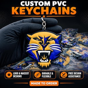 May include: A custom PVC keychain featuring a stylized yellow and blue panther mascot head with white fangs and whiskers. The keychain is attached to a silver metal ring. Text on the image reads CUSTOM PVC KEYCHAINS, LOGO & MASCOT DESIGNS, DURABLE & FLEXIBLE, FREE DESIGN ASSISTANCE, and MADE TO ORDER.