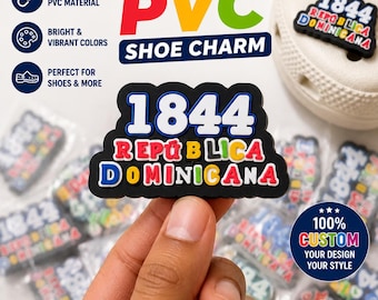 May include: A black PVC shoe charm featuring the white text 1844 above the multi-colored text REPÚBLICA DOMINICANA. The charm has a black background and is displayed alongside other identical charms and a white shoe with holes.