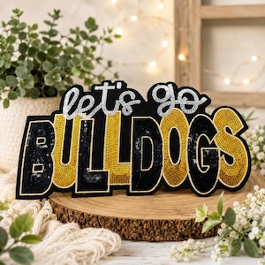 May include: A decorative patch featuring the text let's go in silver sequins and the word BULLDOGS in alternating black and gold sequins. The letters are outlined in a thin gold border, creating a bold, high-contrast design for apparel.