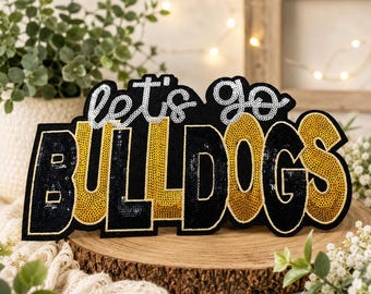 May include: A decorative patch featuring the text let's go in silver sequins and the word BULLDOGS in alternating black and gold sequins. The letters are outlined in a thin gold border, creating a bold, high-contrast design for apparel.