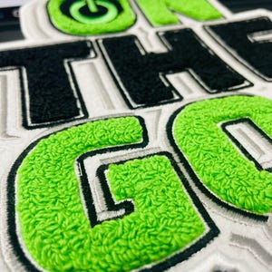 Może przedstawiać: Zbliżenie naszywki z napisem "ON THE GO" w stylu uniwersyteckim. Litery są czarne i zielone z białym obramowaniem. "O" w "ON" ma zielony symbol przycisku zasilania.