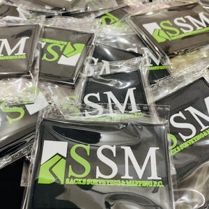 May include: A collection of rectangular black rubber patches individually sealed in clear plastic bags. Each patch features a white and lime green logo with the text SSM and SACKS SURVEYING & MAPPING P.C. printed in white and green.