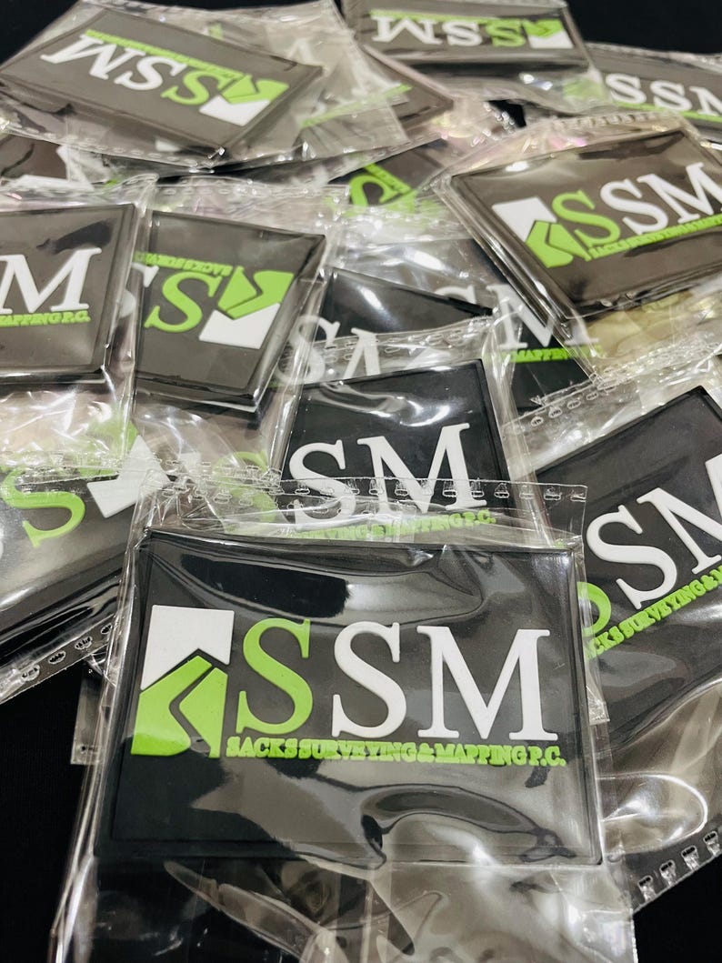May include: A collection of rectangular black rubber patches individually sealed in clear plastic bags. Each patch features a white and lime green logo with the text SSM and SACKS SURVEYING & MAPPING P.C. printed in white and green.