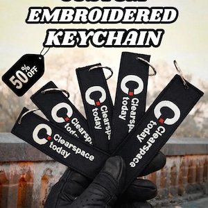 Könnte beinhalten: In einer behandschuhten Hand werden schwarze, bestickte Schlüsselanhänger mit silbernen Ringen gehalten. Jeder Schlüsselanhänger trägt den Text "Clearspace today" und ein weißes "C"-Logo. Ein Etikett zeigt "50% OFF".