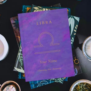May include: A purple Libra journal with gold lettering and a zodiac symbol. The journal has the words "LIBRA", "Your Name", and the phrases "Charming · Fair · Romantic". The journal is stacked on top of other books.