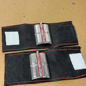 May include: Two black leather wrist cuffs with red stitching and silver and red rectangular accents. Each cuff has a white velcro patch for closure. The cuffs are displayed on a brown surface.
