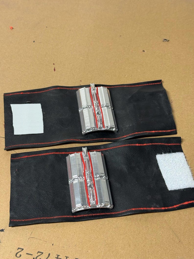 May include: Two black leather wrist cuffs with red stitching and silver and red rectangular accents. Each cuff has a white velcro patch for closure. The cuffs are displayed on a brown surface.