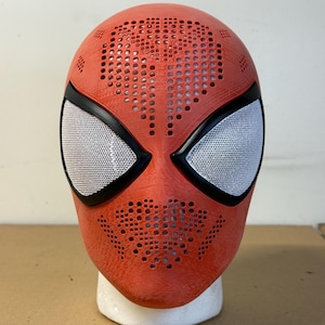 May include: A red Spider-Man mask with white mesh eye coverings and black trim. The mask has a textured surface and numerous small holes across the top and face. The mask is displayed on a white foam head.